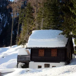 Ein kleines Holzhaus mit schneebedecktem Dach steht in einer verschneiten Landschaft umgeben von Kiefern. Das Haus befindet sich in einem Berggebiet mit Schneehaufen darum herum.