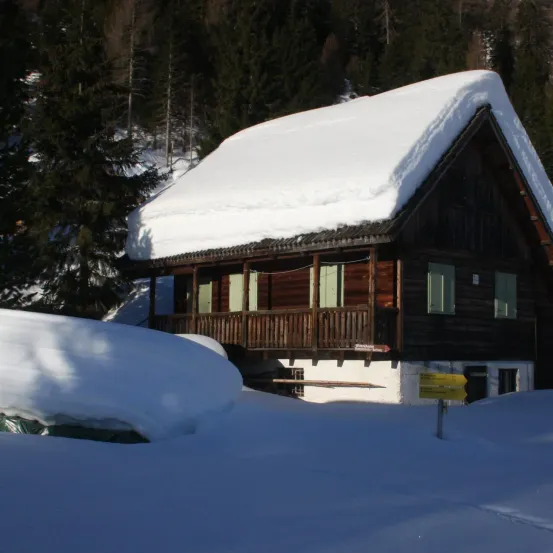 Ein Holzhaus mit schneebedecktem Dach, umgeben von Schnee in einem Wald. Bäume sind im Hintergrund und ein gelbes Schild ist im Vordergrund.