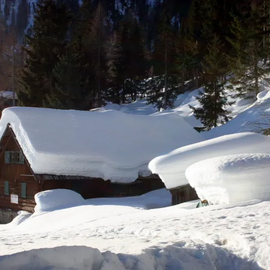 Ein verschneiter Holzchalet in einem verschneiten Gebirgsgebiet mit Bäumen und Hügeln im Hintergrund.