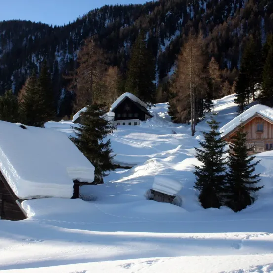Schneebedeckte Hütten in einer bergigen Landschaft mit Bäumen und einem hellen Himmel. Die Hütten sind von Schnee umgeben, und die Bäume sind ebenfalls schneebedeckt.