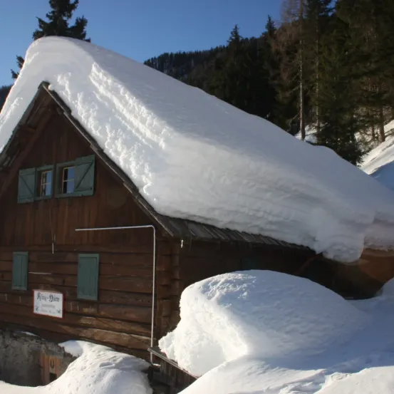Ein holzverkleidetes Haus, das mit Schnee bedeckt ist, hat grüne Fensterläden und ein Schild mit der Aufschrift 'Kina-Datte' davor. Bäume umgeben das Haus.