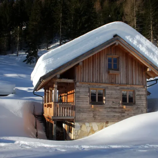 Ein von Schnee umgebenes Holzchalet mit einer Person auf dem Balkon in einer verschneiten Landschaft mit Bäumen im Hintergrund.