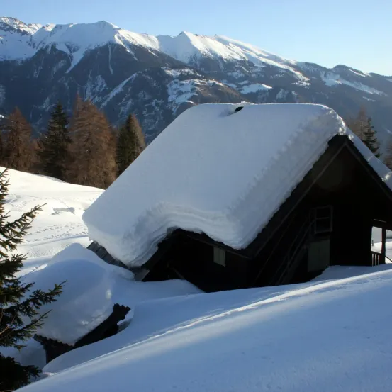 Ein kleines Chalet, das in einer verschneiten Bergregion mit immergrünen Bäumen und schneebedeckten Bergen im Hintergrund von Schnee bedeckt ist.