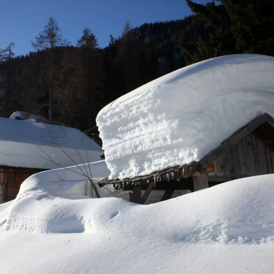 Eine verschneite Berglandschaft mit einem schneebedeckten Haus, Bäumen und einem klaren blauen Himmel.