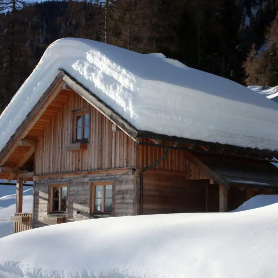 Ein Holzchalet mit einer dicken Schneeschicht auf dem Dach, eingebettet in eine verschneite Landschaft mit Bäumen im Hintergrund.