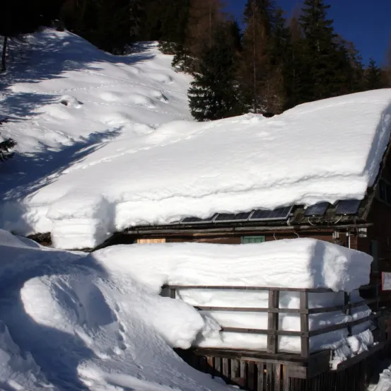 Eine Holzhütte mit schneebedecktem Dach und Solarmodulen, umgeben von schneebedeckter Landschaft und Bäumen.