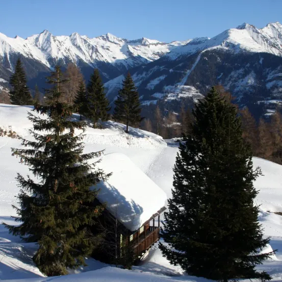 Ein kleines Holzchalet, das mit Schnee bedeckt ist, mit Tannen in einer verschneiten Landschaft mit Bergen im Hintergrund.