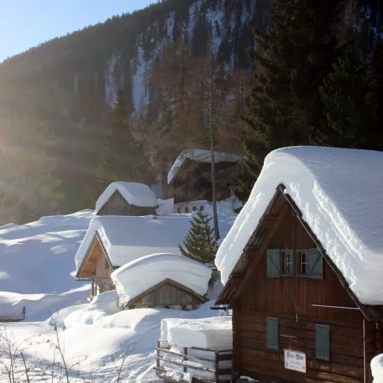 Eine schneebedeckte Landschaft mit mehreren Holzhütten, darunter eine mit einem Schild, unter einem hellen Himmel mit einem Berg im Hintergrund.