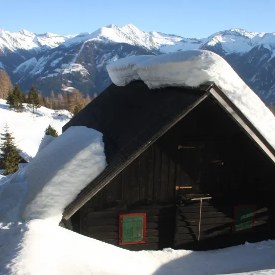 Eine kleine Hütte ist unter dickem Schnee vergraben, in einer verschneiten Berglandschaft mit immergrünen Bäumen. Das Dach ist geneigt, und eine Schaufel lehnt an der Wand.