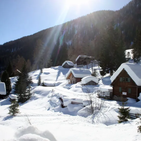 Eine verschneite Landschaft mit einem Bergrücken im Hintergrund und mehreren kleinen Holzhütten, die mit Schnee bedeckt sind. Die Sonne scheint hell und wirft ein warmes Licht auf den Schnee.