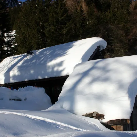 Ein schneebedecktes Holzhaus steht in einer verschneiten Landschaft, mit Bäumen im Hintergrund. Das Dach und der Boden sind mit Schnee bedeckt.