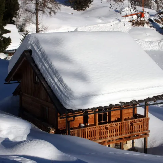 Ein kleines Holzchalet, bedeckt mit Schnee, steht in einer verschneiten Landschaft, umgeben von Bäumen und einer entfernten Skiliftstation.