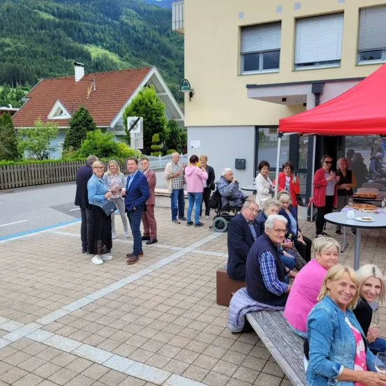 Mehrere Menschen sind im Freien versammelt, möglicherweise für eine Gemeinschaftsveranstaltung. Einige stehen, während andere auf einer Bank unter einem roten Zelt sitzen. In der Nähe befindet sich ein Gebäude mit einem roten Dach und einem Tisch mit Essen.