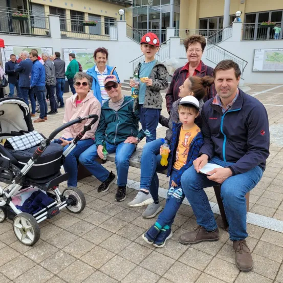 Eine Gruppe von Menschen sitzt auf einer Bank im Freien mit einem Gebäude und Treppen hinter ihnen. Einige halten Essen und Getränke. Ein Baby im Kinderwagen und ein junger Junge im Spider-Man-Hut sind ebenfalls anwesend.