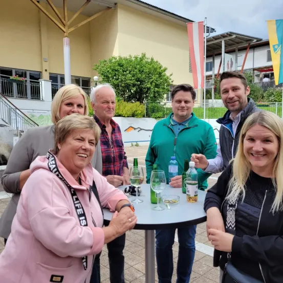 Eine Gruppe lächelnder Erwachsener, zwei Frauen und vier Männer, stehen um einen Tisch mit Getränken, möglicherweise bei einer sozialen Veranstaltung. Sie scheinen für ein Foto zu posieren. Dahinter ist ein Gebäude mit Flaggen und Grünpflanzen zu sehen.