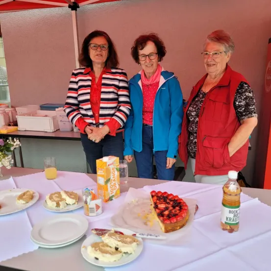 Drei Frauen stehen hinter einem Tisch mit verschiedenen Lebensmitteln. Eine Frau trägt ein gestreiftes Shirt, eine andere eine blaue Jacke und die dritte eine rote Weste. Eine Flasche Saft, ein Kuchen und eine Packung Orangensaft stehen auf dem Tisch.