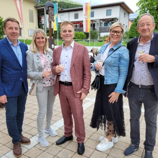 Fünf Personen posieren für ein Foto auf einer gepflasterten Fläche, jeweils mit einem Glas Wein in der Hand. Die Gruppe besteht aus zwei Männern und drei Frauen. Hinter ihnen befindet sich eine Kulisse aus Gebäuden und Bäumen.