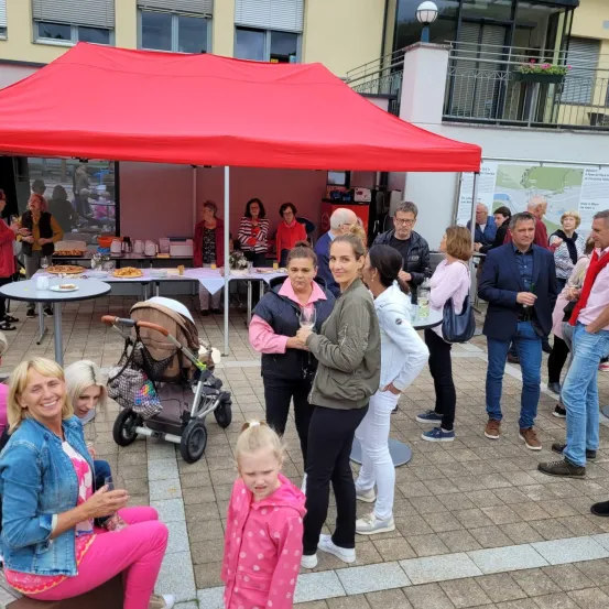 Eine Gruppe von Menschen versammelt sich unter einem roten Zelt und genießt Essen und Getränke. Ein Kinderwagen steht in der Nähe, und ein junges Mädchen in einer rosa Jacke steht zwischen zwei Frauen.