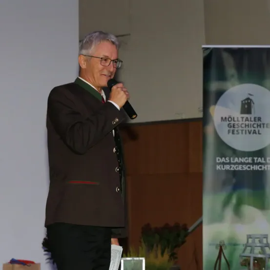 Ein älterer Mann spricht in ein Mikrofon bei einer Veranstaltung. Hinter ihm ist ein Banner für das Mölltaler Geschichtsfestival zu sehen.
