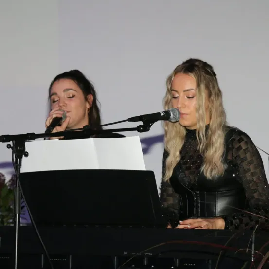 Zwei Frauen treten auf der Bühne auf, eine singt in ein Mikrofon, während die andere von einem Notenständer liest. Sie sind in Schwarz gekleidet und vor einem Klavier positioniert.