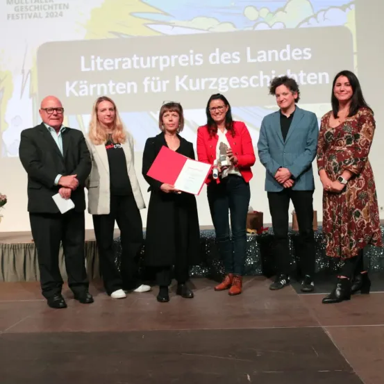 Eine Gruppe von Menschen steht lächelnd auf der Bühne, einer hält ein Zertifikat und einen Pokal. Ein Banner hinter ihnen trägt die Aufschrift 'Literaturpreis des Landes Kärnten für Kurzgeschichten'.