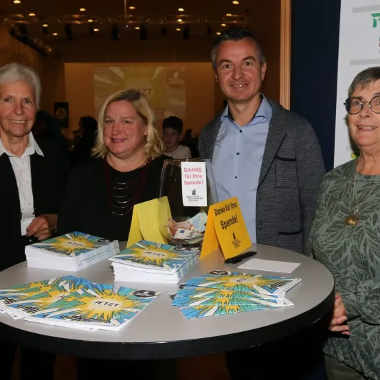 Vier Personen stehen um einen Tisch mit verschiedenen Materialien. Ein Glas mit Geld und eine Notiz Danke fur Ihre Spende! steht auf dem Tisch. Hinter ihnen zeigt eine Projektionsleinwand eine Präsentation.