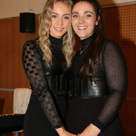 Zwei Frauen in schwarzen Outfits stehen eng beieinander und lächeln für ein Foto. Eine hat lange Haare, die andere trägt eine Halskette. Hinter ihnen ist eine Holzwand und ein schwarzer Lautsprecher.