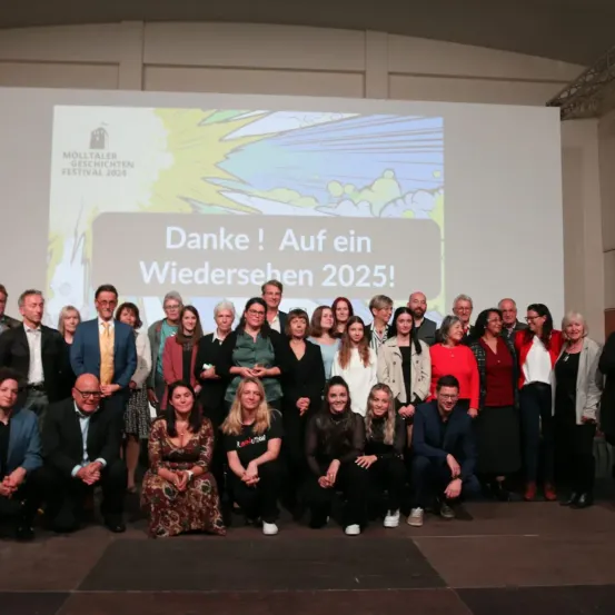 Eine Gruppe von Menschen posiert für ein Foto auf der Bühne vor einer Leinwand mit der Aufschrift 'Danke! Auf ein Wiedersehen 2025!' Die Teilnehmer sind feierlich gekleidet.