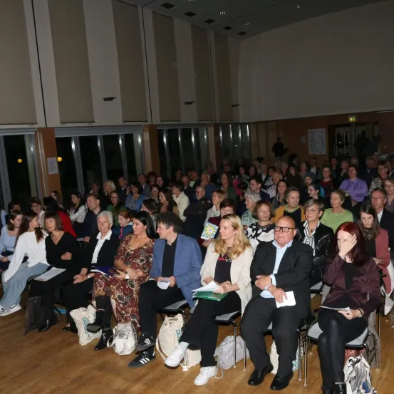 Eine große Gruppe von Menschen sitzt in Reihen in einem Raum, einige lesen, andere lauschen aufmerksam. Der Raum hat Holzböden und Glaswände.