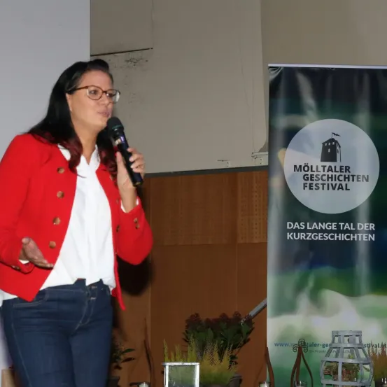 Eine Frau in einer roten Jacke spricht in ein Mikrofon bei einer Konferenz. Ein Banner mit den Worten 'Mölltaler Geschichten Festival' ist hinter ihr sichtbar.