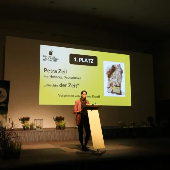 Eine Frau steht auf der Bühne vor einem Rednerpult mit Mikrofon und präsentiert. Hinter ihr ist eine Leinwand mit dem Titel "1. Platz" und einem Bild von Petra Zeil.