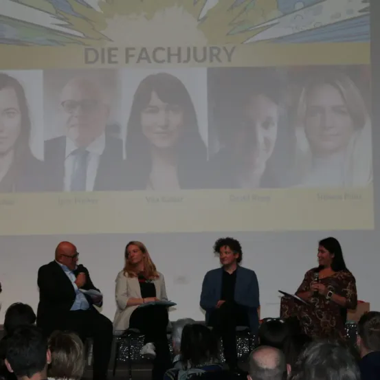 Eine Podiumsdiskussion mit vier Personen, zwei Männern und zwei Frauen, findet vor einem Publikum statt. Ein großer Bildschirm hinter ihnen zeigt den Text 'DIE FACHJURY' mit Bildern von vier Personen.