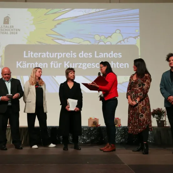 Fünf Personen stehen auf einer Bühne, eine hält ein Mikrofon und liest aus einer Mappe. Hinter ihnen ist eine große Projektionsleinwand mit einer Präsentation über den Literaturpreis des Landes Kärnten für Kurzgeschichten.