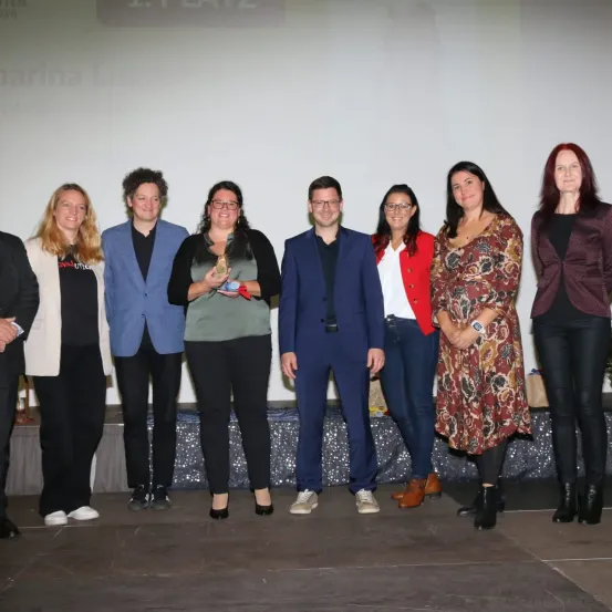 Eine Gruppe von Menschen steht gemeinsam auf der Bühne, lächelt und posiert für ein Foto. Eine Frau hält eine Trophäe und ein Mann steht neben ihr. Hinter ihnen zeigt eine Leinwand den Text '1. PLATZ'. Sie befinden sich wahrscheinlich bei einer Preisverleihung.