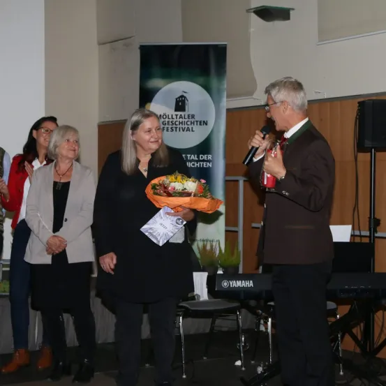 Eine Preisverleihung, bei der eine Frau in einem schwarzen Kleid einen Blumenstrauß hält und lächelt. Hinter ihr steht ein Mann mit einem Mikrofon. Auf der linken Seite beobachten zwei Frauen in Mänteln. Ein Banner mit 'Mölltaler Geschichten Festival' hängt im Hintergrund.