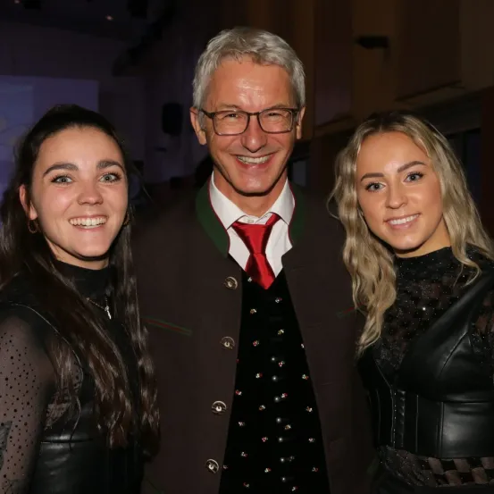 Ein Mann in traditioneller bayerischer Kleidung steht zwischen zwei Frauen und lächelt für ein Foto. Der Mann trägt eine Brille und eine rote Krawatte. Die Frauen haben blonde und dunkle Haare.