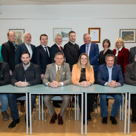 Eine Gruppe formell gekleideter Personen, darunter Männer und Frauen, sitzen um Tische herum in einer Besprechung. Sie scheinen sich in einer professionellen Umgebung zu befinden, möglicherweise auf einer Konferenz oder einem Workshop.