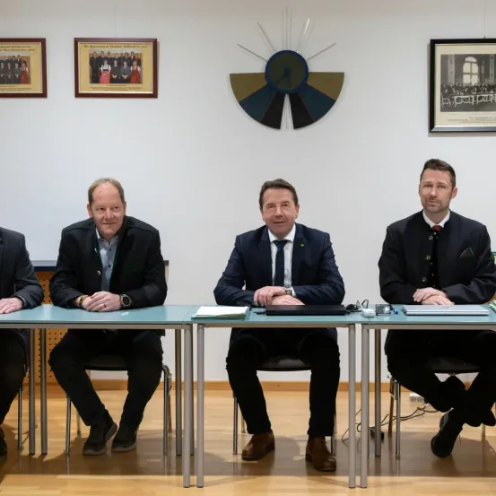 Vier Männer in Anzügen sitzen an einem Tisch, wahrscheinlich in einem Konferenzraum. Der Mann rechts hat die Füße auf dem Tisch.