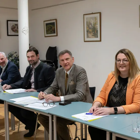 Vier Personen sitzen an einem Tisch in einem Besprechungsraum mit Papieren und einem Stift. Eine Frau trägt eine Brille und eine Uhr. Ein Mann neben ihr hält eine Brille und einen Stift. Hinter ihnen hängen gerahmte Bilder an der Wand.