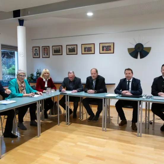 Eine Besprechung mit sechs Personen an einem Tisch. Einer schreibt, während andere sich unterhalten. Hinter ihnen schmücken Bilderrahmen die Wand.