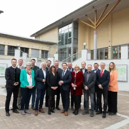 Eine Gruppe von formell gekleideten Personen steht vor einem modernen Gebäude mit Glaswänden und einer Treppe. Sie posieren für ein Foto.