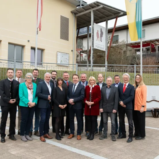 Eine Gruppe formell gekleideter Personen, sowohl Männer als auch Frauen, stehen vor einem Gebäude mit Fahnen und einem Schild, das Kindergarten Muhldorf besagt.