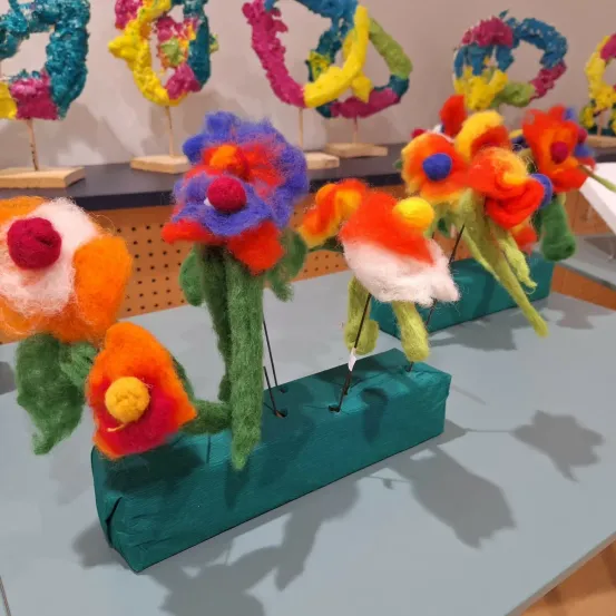 Mehrere bunte Filzblumen sind auf einer grünen Basis ausgestellt. Dahinter befinden sich weitere bunte Filzblumen auf Holzständern vor einem weißen Hintergrund.