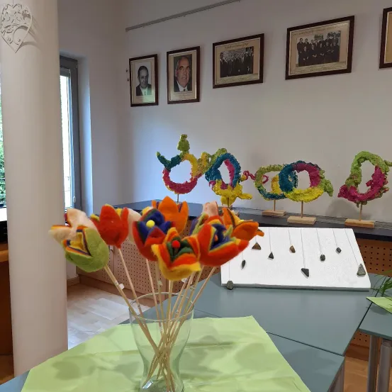 Ein Raum zeigt farbenfrohe, handgefertigte Blumen auf einem Tisch, mit gerahmten Bildern an der Wand und einer Ausstellung von Steinen auf einem weißen Brett.