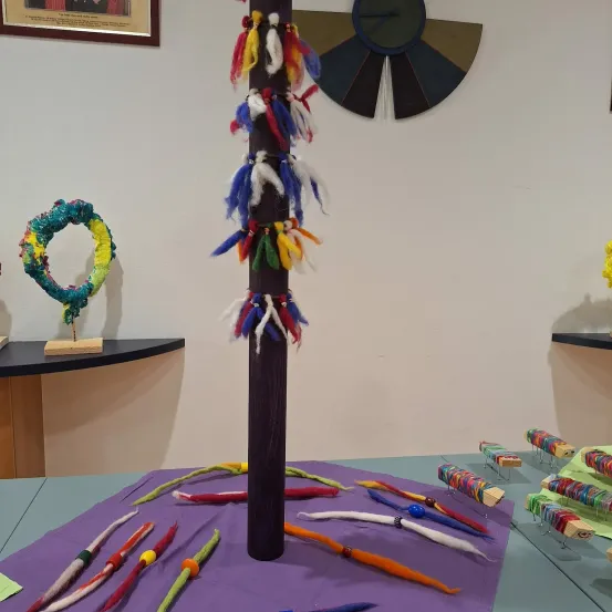 Eine Ausstellung farbenfroher handgefertigter Gegenstände auf einem Tisch mit einem hohen, mit Federn verzierten Pfosten. An der Wand befindet sich ein gerahmtes Bild und ein rundes, segmentiertes Kunstwerk.