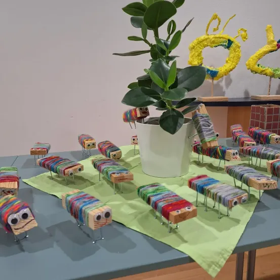 Ein Tisch zeigt eine grüne Pflanze in einem weißen Topf, umgeben von bunten hölzernen Raupen mit Garn und Augen. Dahinter stehen zwei gelbe Papierschmetterlinge auf einem Regal mit einer Ziegelwand im Hintergrund.