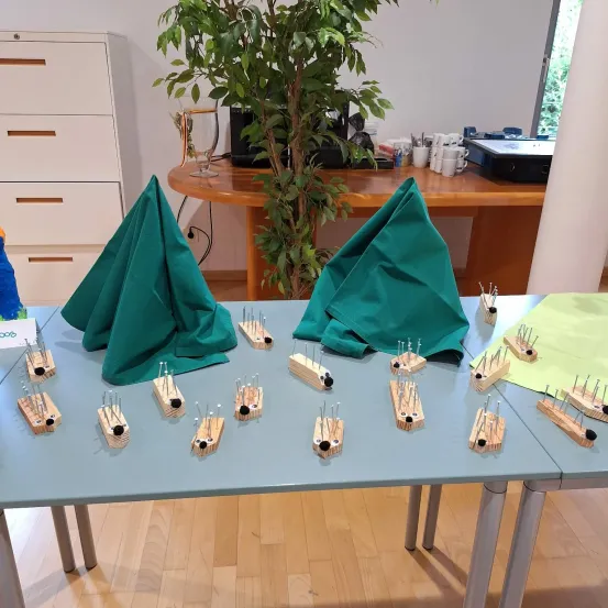 Ein Tisch mit mehreren Holzobjekten mit Stecknadeln darauf und grünen Tüchern obenauf, eine Pflanze und ein Schreibtisch im Hintergrund.