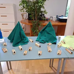 Ein Tisch mit mehreren Holzobjekten mit Stecknadeln darauf und grünen Tüchern obenauf, eine Pflanze und ein Schreibtisch im Hintergrund.