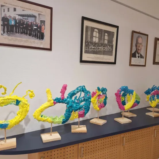 Eine Wand mit drei gerahmten Bildern und einem Regal mit farbenfrohen Skulpturen darauf. Die Bilder zeigen ein Gruppenfoto, eine formelle Versammlung und ein Einzelporträt.