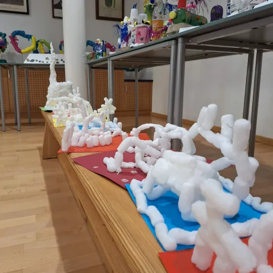 Ein langer Holztisch zeigt verschiedene Skulpturen aus weißer Baumwolle, die in einer Reihe auf einem blauen und roten Papieruntergrund angeordnet sind. Hinter dem Tisch steht ein Metalltisch mit bunten, verspielten Objekten.